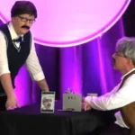 Jason and Kristin on stage with wigs and false moustaches have a corporate discussion